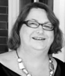 A black and white head shot of Paula Hanlon. She is smiling, wears glasses and a necklace.