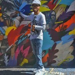 A colour photograph of Nolan Stevens standing near a wall covered in street art. He is wearing a light-blue peak cap and pointing to the wall.