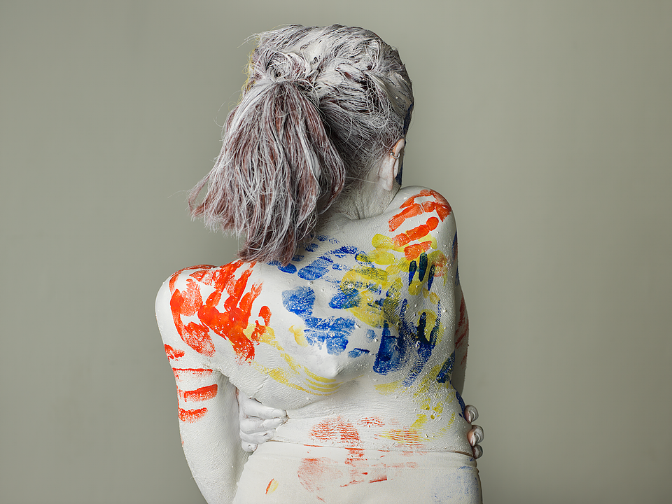 Becky Dann is facing the wall. Her body is painted white and she has crossed her arms. The tips of her fingers are visible holding her waist. There are red, blue and yellow hand prints over her back.